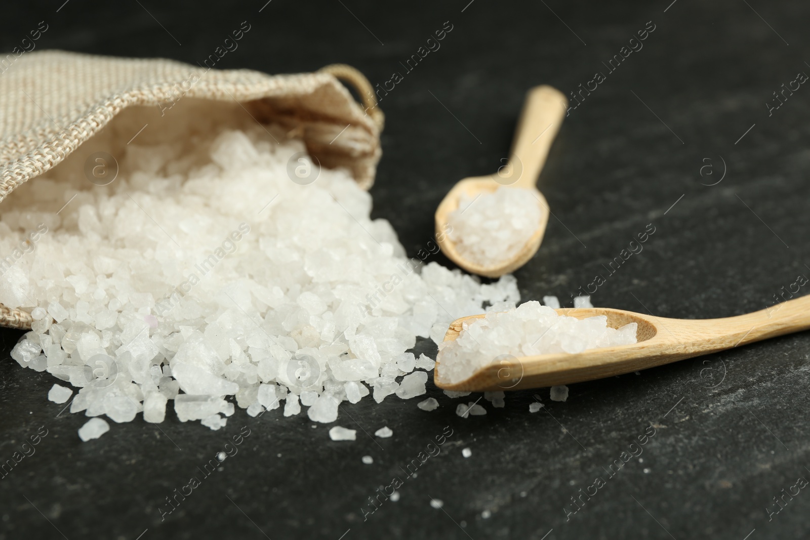 Photo of Sea salt, bag and spoons on dark gray textured table, closeup