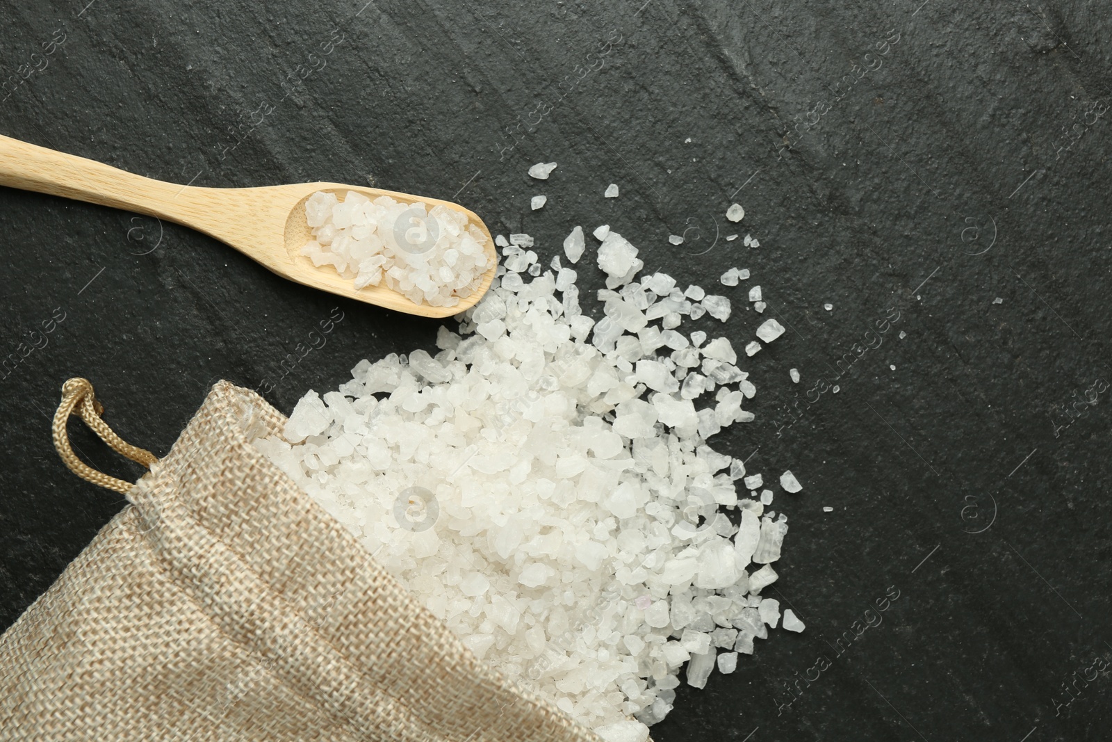 Photo of Sea salt, bag and spoon on dark gray textured table, flat lay