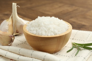 Photo of Sea salt in bowl, rosemary and garlic on brown table, closeup