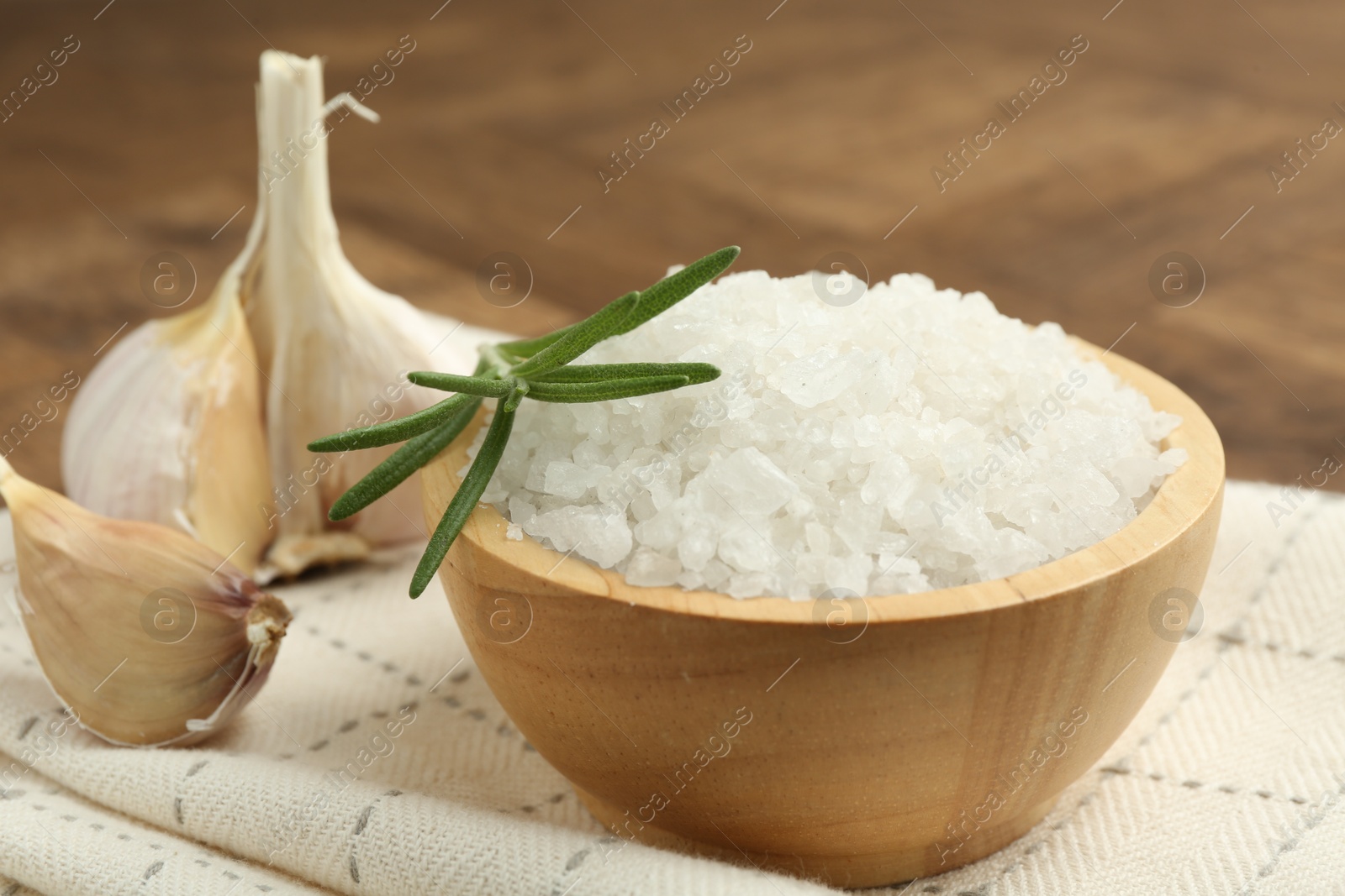 Photo of Sea salt in bowl, rosemary and garlic on brown table, closeup