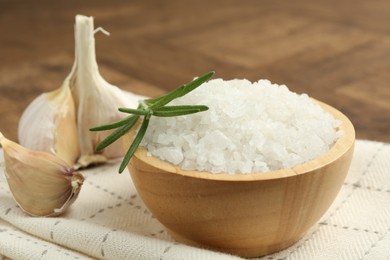 Photo of Sea salt in bowl, rosemary and garlic on brown table, closeup