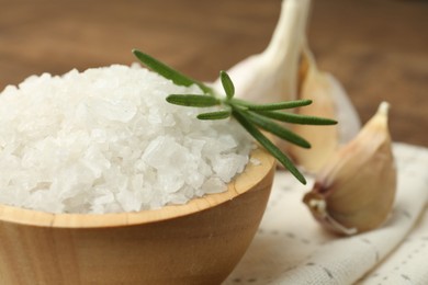 Photo of Sea salt in bowl, rosemary and garlic on brown table, closeup