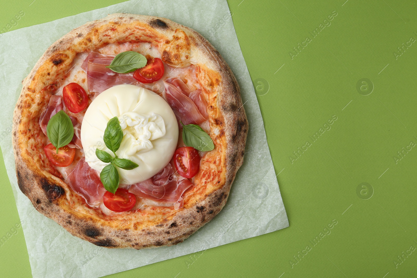 Photo of Delicious pizza with burrata cheese, basil, tomato and ham on green background, top view. Space for text