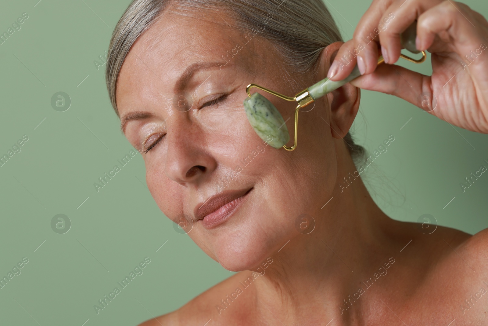 Photo of Beautiful woman doing facial massage with roller on light green background, closeup