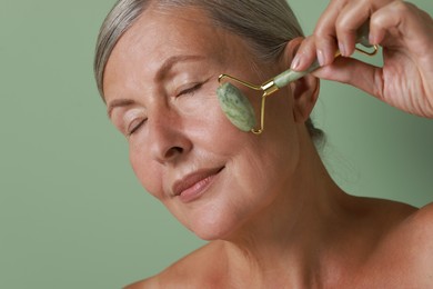 Beautiful woman doing facial massage with roller on light green background, closeup