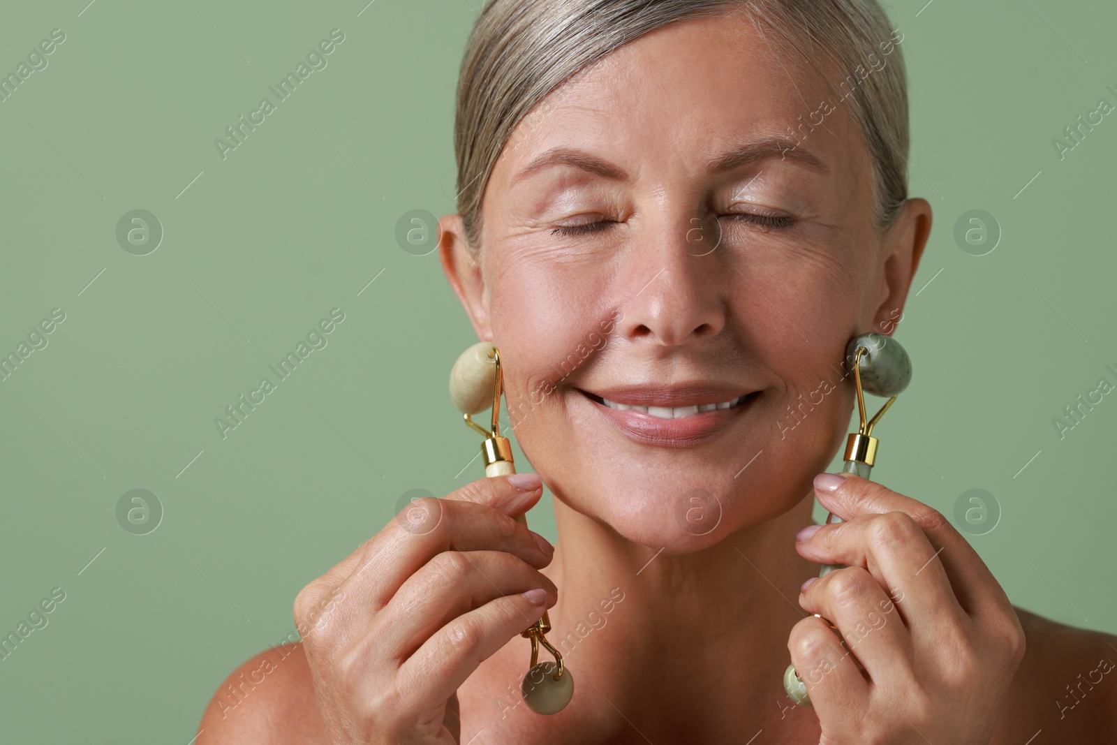 Photo of Beautiful woman doing facial massage with rollers on light green background, closeup