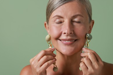 Photo of Beautiful woman doing facial massage with rollers on light green background, closeup