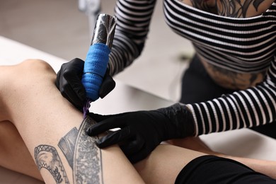 Photo of Professional artist tattooing woman's leg with machine in salon, closeup