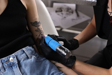 Professional artist tattooing woman's arm with machine in salon, closeup