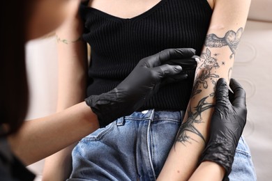 Professional artist in gloves applying cream onto woman's arm with fresh tattoo in salon, closeup