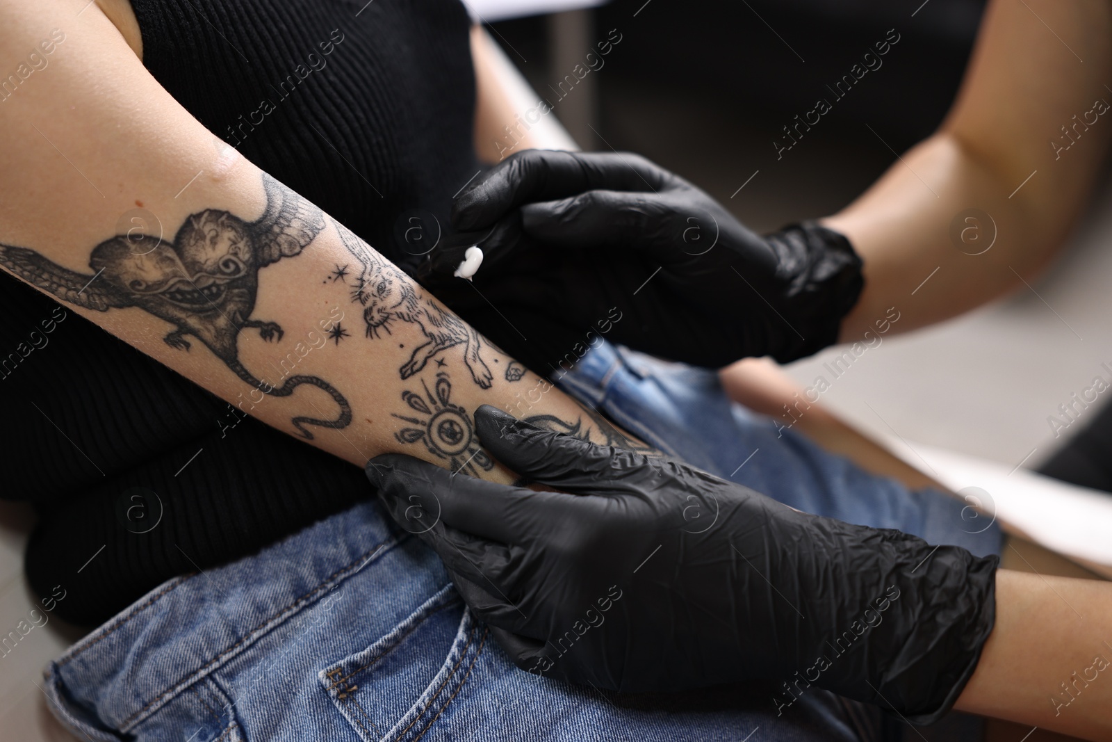 Photo of Professional artist in gloves applying cream onto woman's arm with fresh tattoo in salon, closeup