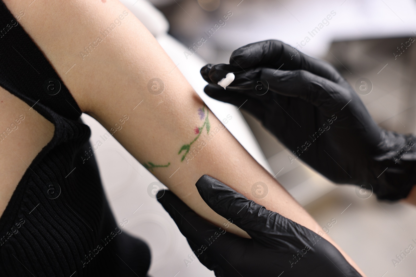 Photo of Professional artist in gloves applying cream onto woman's arm with fresh tattoo in salon, closeup