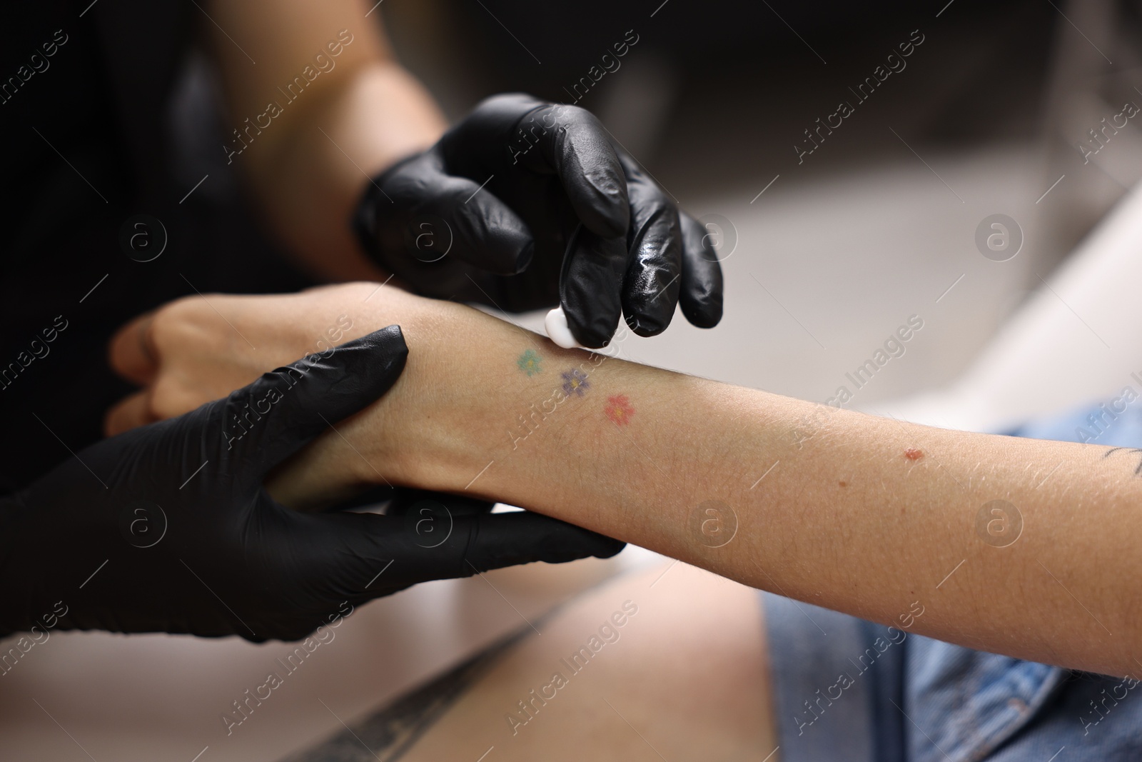 Photo of Professional artist in gloves applying cream onto woman's hand with fresh tattoo in salon, closeup