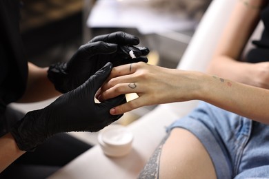 Professional artist in gloves applying cream onto woman's finger with fresh tattoo in salon, closeup