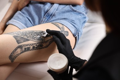 Photo of Professional artist in gloves applying cream onto woman's leg with fresh tattoo in salon, closeup