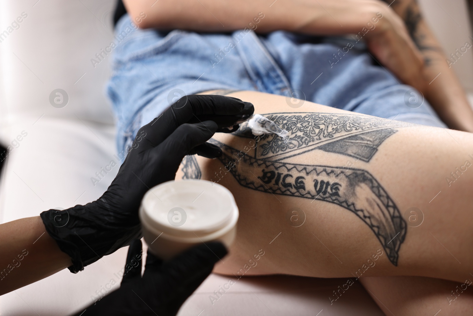 Photo of Professional artist in gloves applying cream onto woman's leg with fresh tattoo in salon, closeup