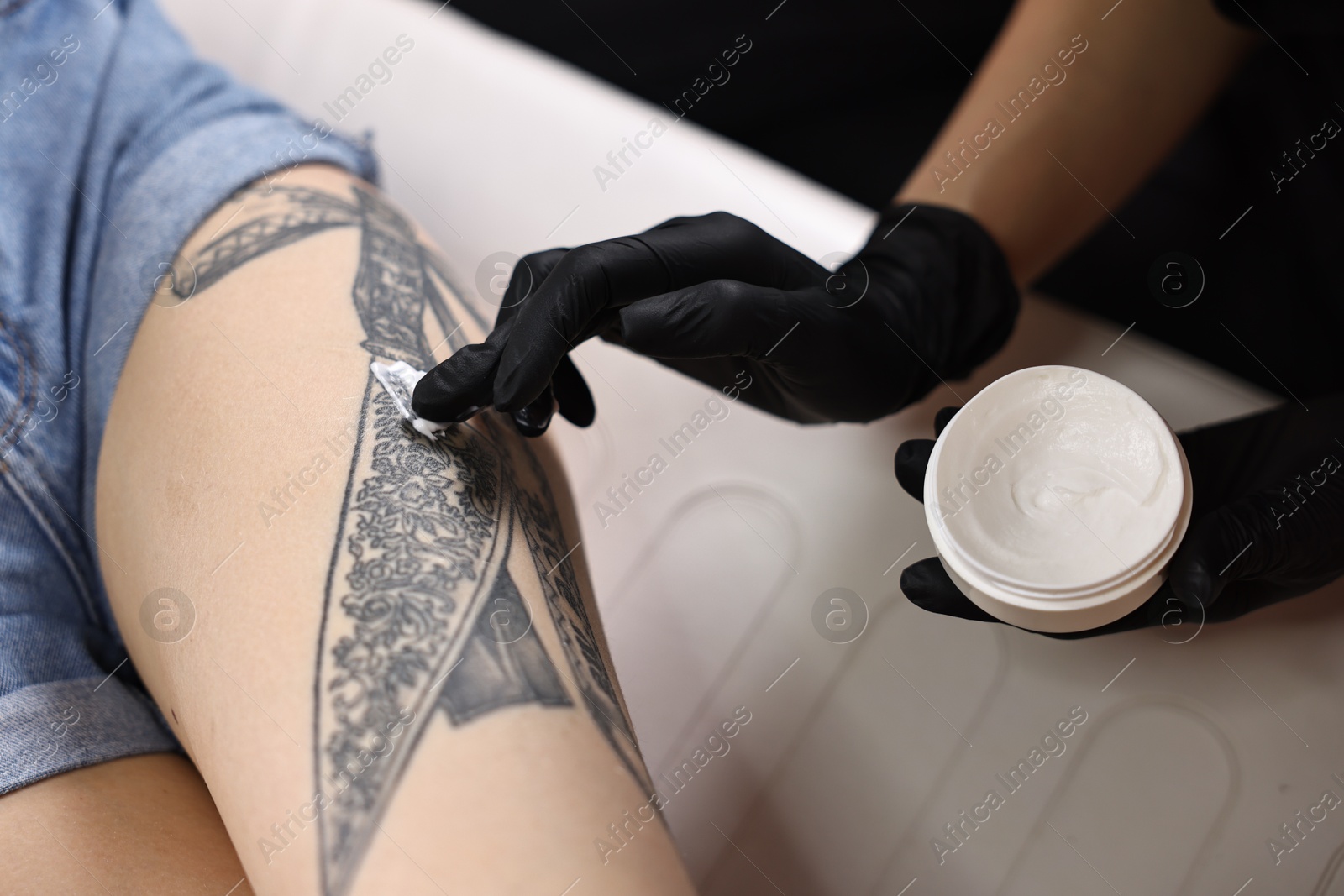 Photo of Professional artist in gloves applying cream onto woman's leg with fresh tattoo in salon, closeup