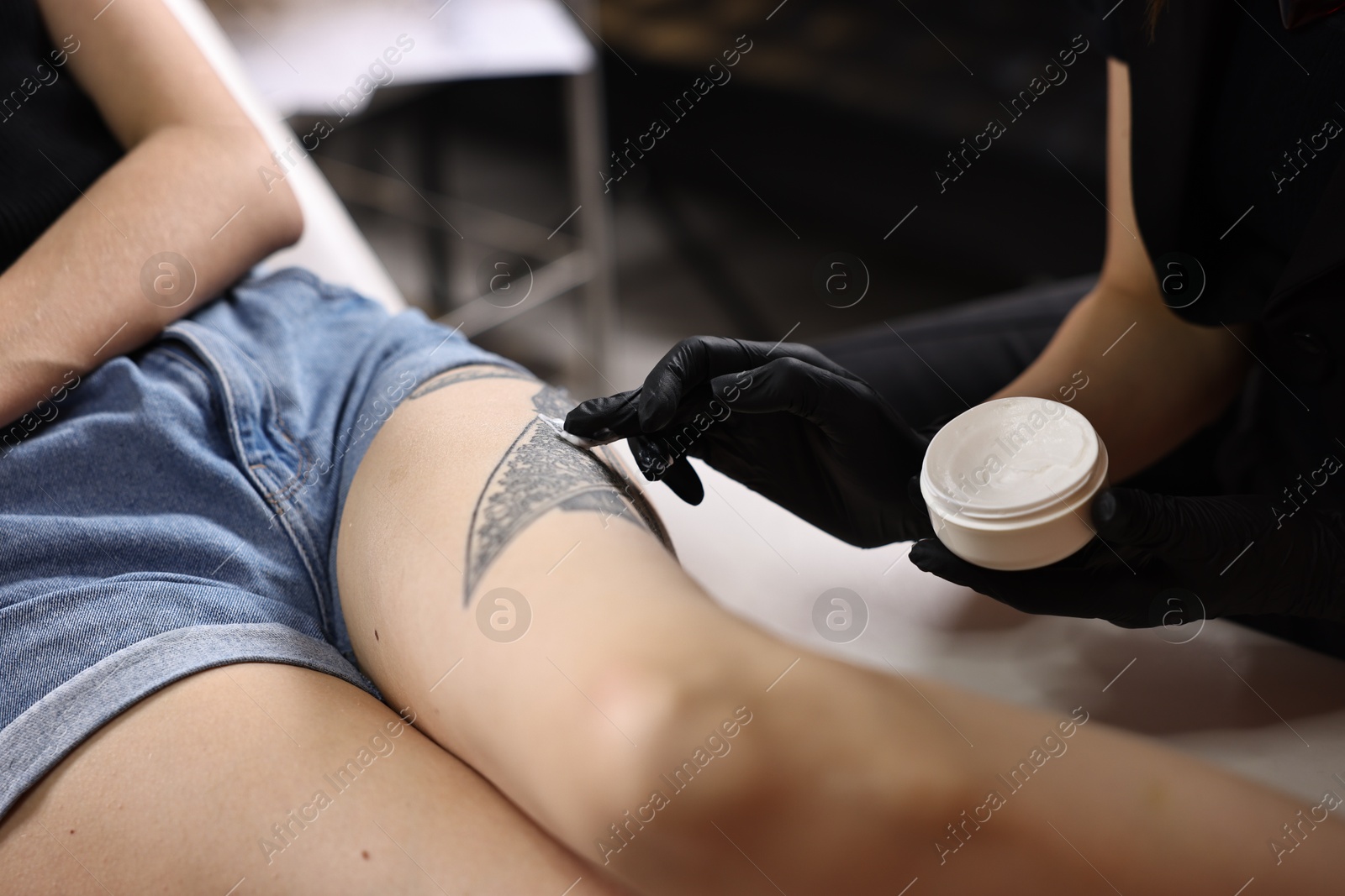 Photo of Professional artist in gloves applying cream onto woman's leg with fresh tattoo in salon, closeup