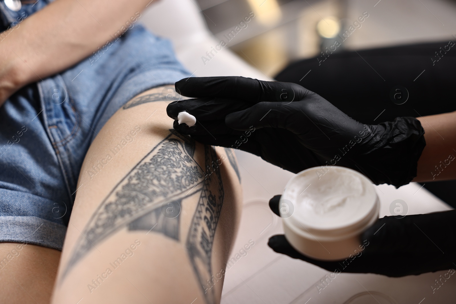 Photo of Professional artist in gloves applying cream onto woman's leg with fresh tattoo in salon, closeup