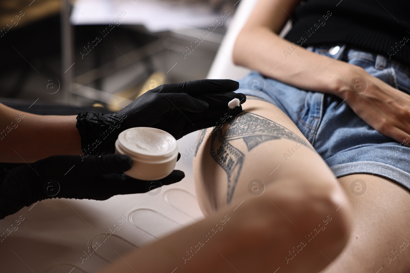 Photo of Professional artist in gloves applying cream onto woman's leg with fresh tattoo in salon, closeup