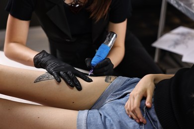 Photo of Professional artist tattooing woman's leg with machine in salon, closeup