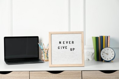 Letter board with phrase Never Give Up, laptop, alarm clock and stationery on white desk