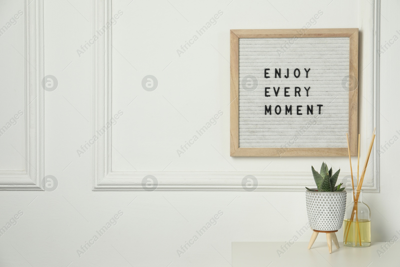 Photo of Letter board with phrase Enjoy Every Moment , reed diffuser and plant on white desk. Space for text