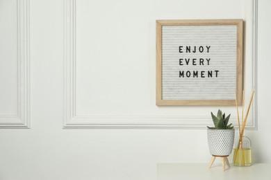 Letter board with phrase Enjoy Every Moment , reed diffuser and plant on white desk. Space for text