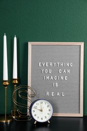 Photo of Letter board with phrase Everything You Can Imagine Is Real, alarm clock, candles and decor element on table near green wall