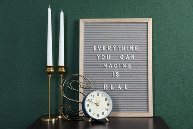 Photo of Letter board with phrase Everything You Can Imagine Is Real, alarm clock, candles and decor element on table near green wall