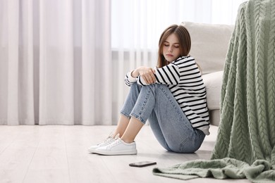 Loneliness concept. Sad teenage girl with smartphone on floor at home, space for text