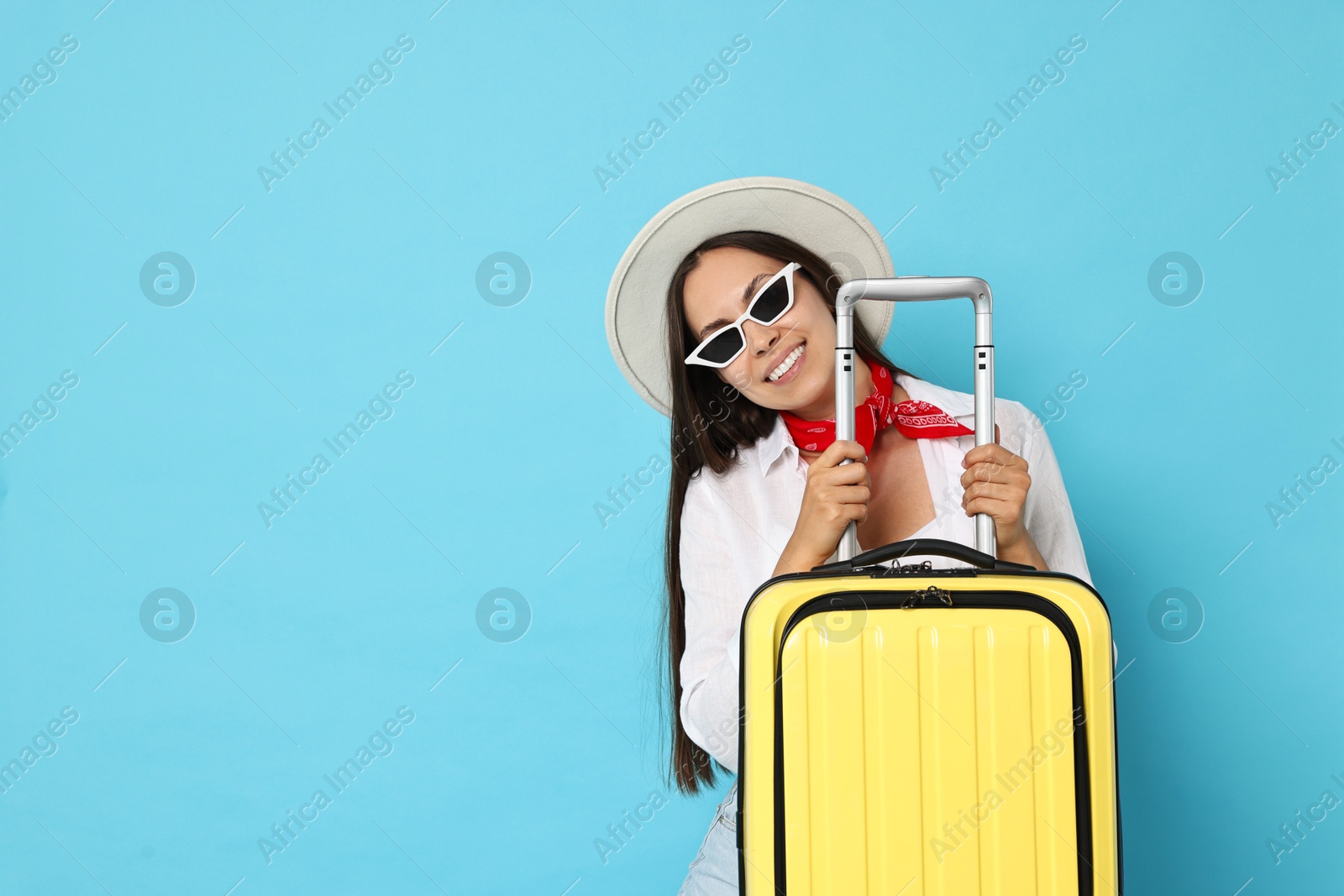 Photo of Happy young woman in sunglasses with suitcase and on light blue background, space for text