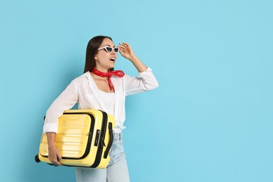 Surprised woman in sunglasses with suitcase on light blue background, space for text