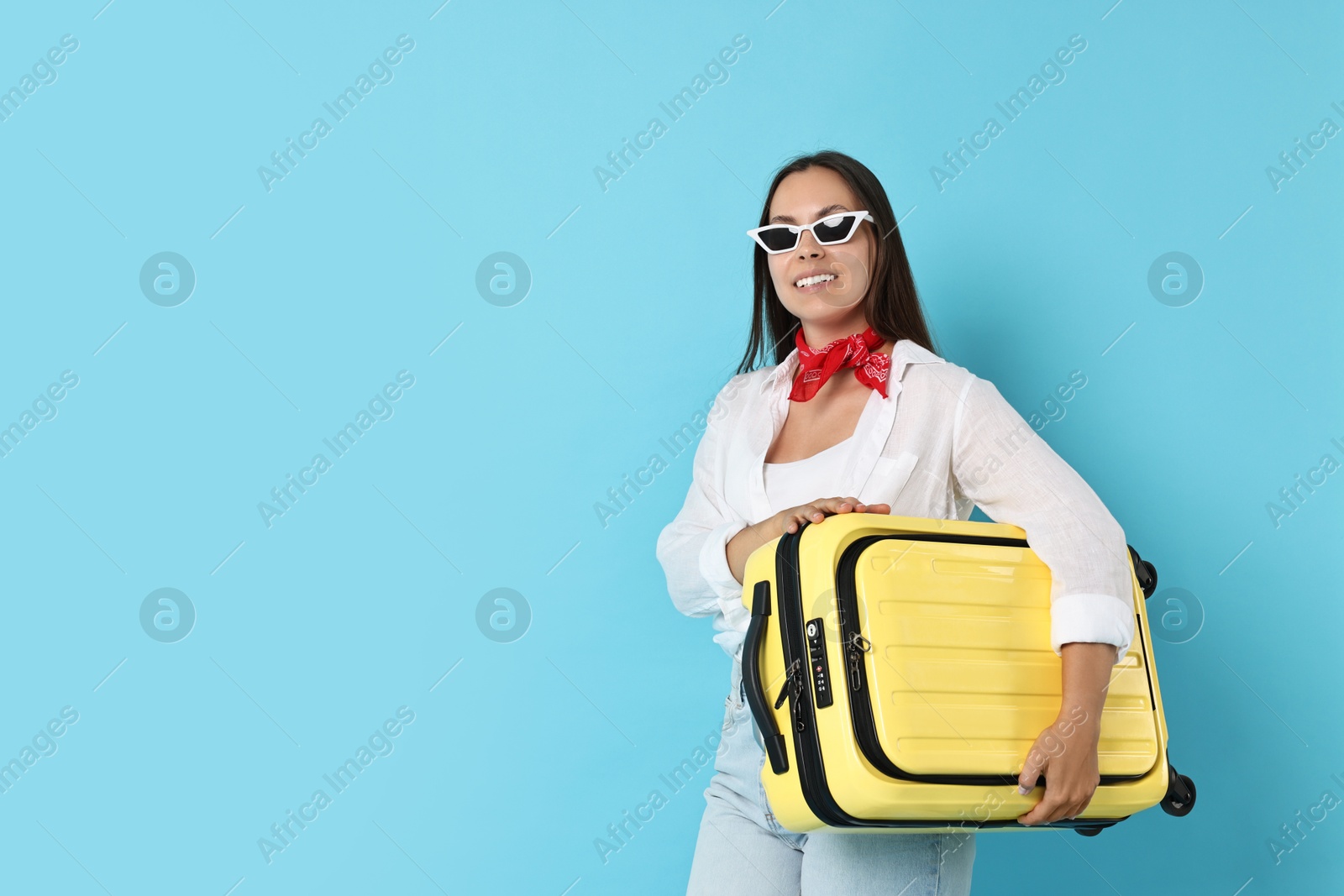 Photo of Happy young woman in sunglasses with suitcase on light blue background, space for text