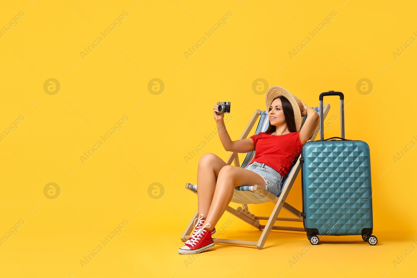 Photo of Happy young woman taking picture while sitting on folding chair and suitcase against orange background, space for text