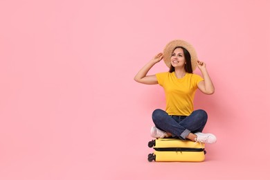 Happy young woman sitting on suitcase against pink background, space for text