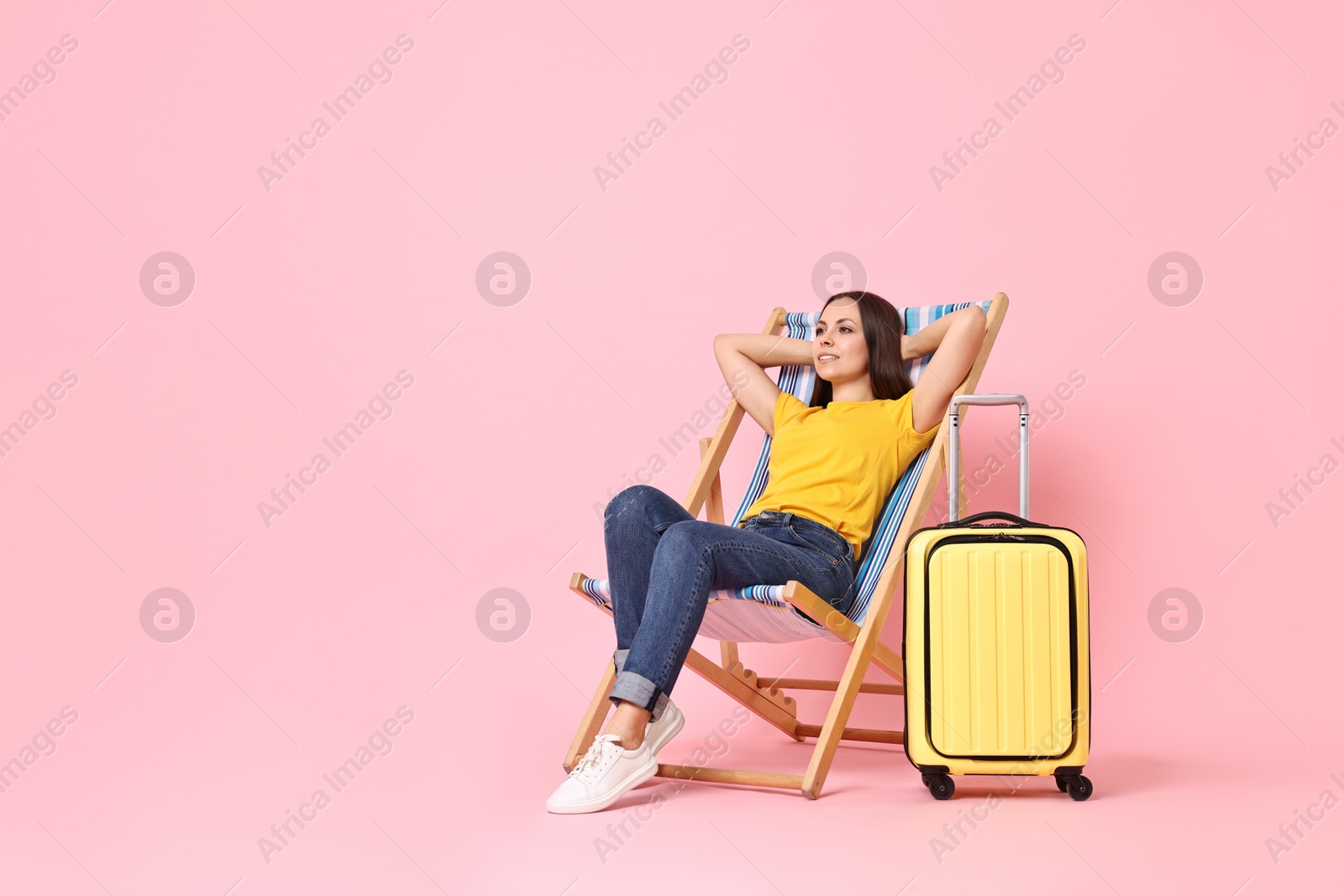 Photo of Happy young woman sitting on folding chair and suitcase against pink background, space for text