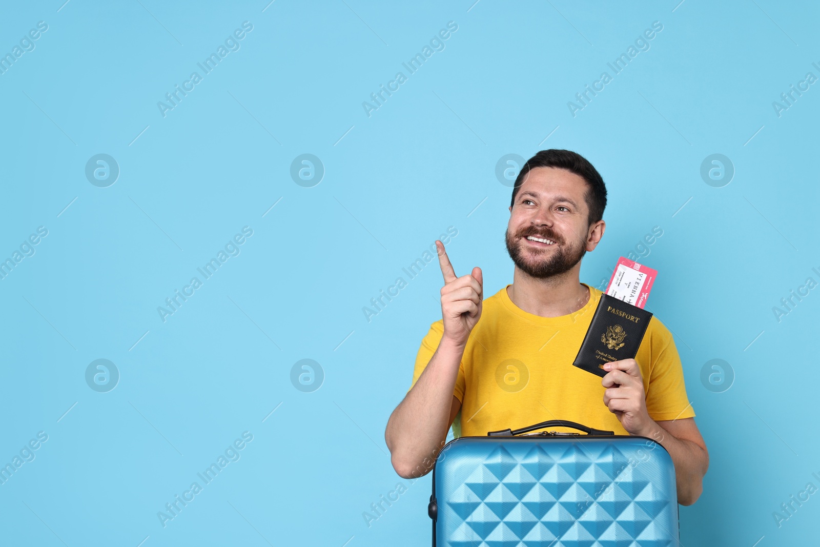 Photo of Happy man with suitcase, passport and ticket pointing at something on light blue background, space for text