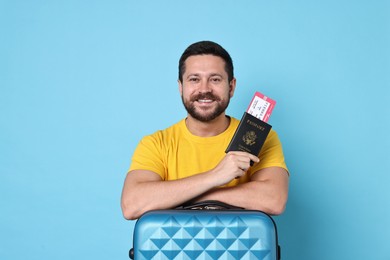 Happy man with suitcase, passport and ticket on light blue background