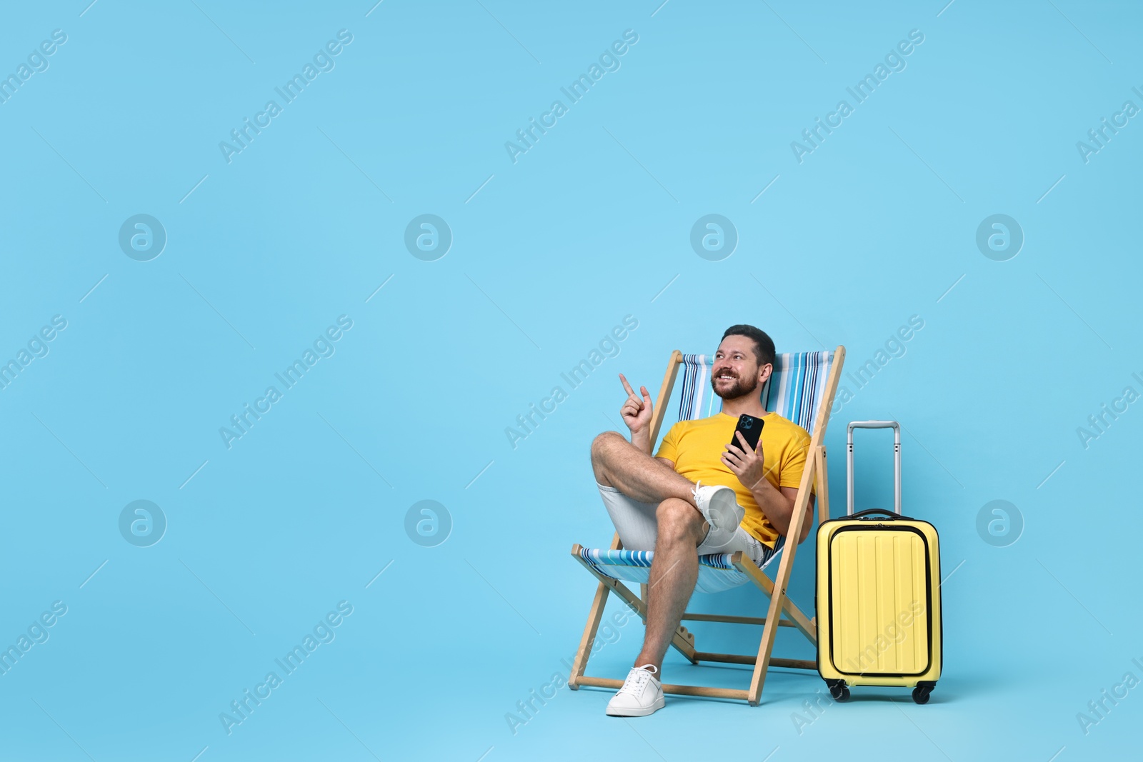 Photo of Happy man with smartphone sitting on folding chair and suitcase against light blue background, space for text