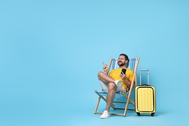 Happy man with smartphone sitting on folding chair and suitcase against light blue background, space for text