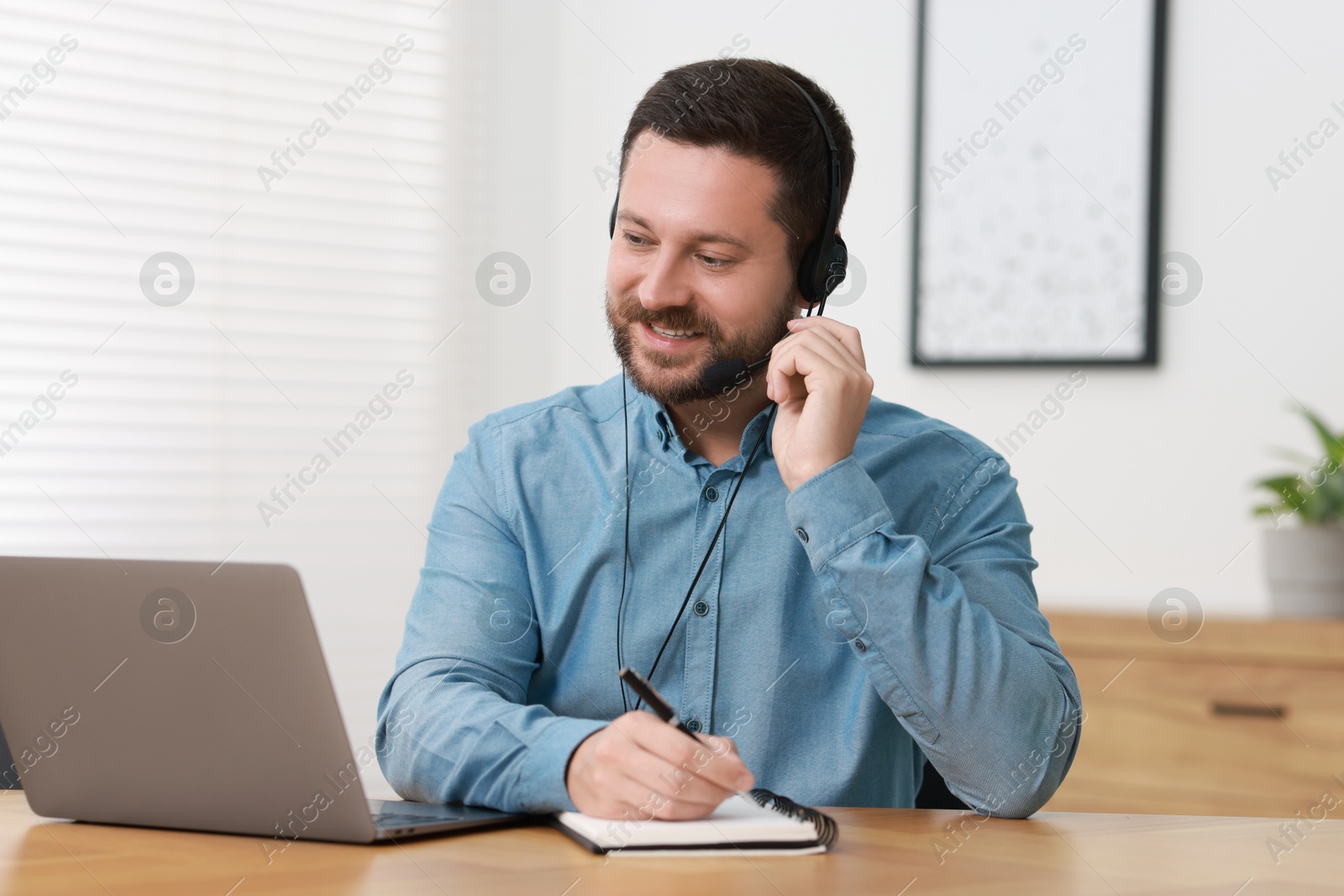 Photo of Interpreter in headset taking notes while having video chat via laptop at wooden table indoors