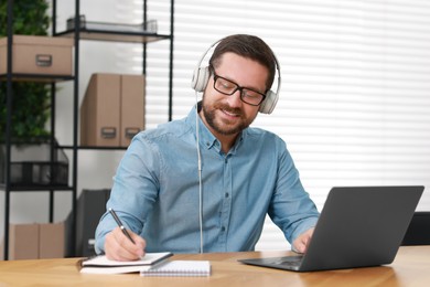 Interpreter in headphones taking notes while having video chat via laptop at wooden table indoors