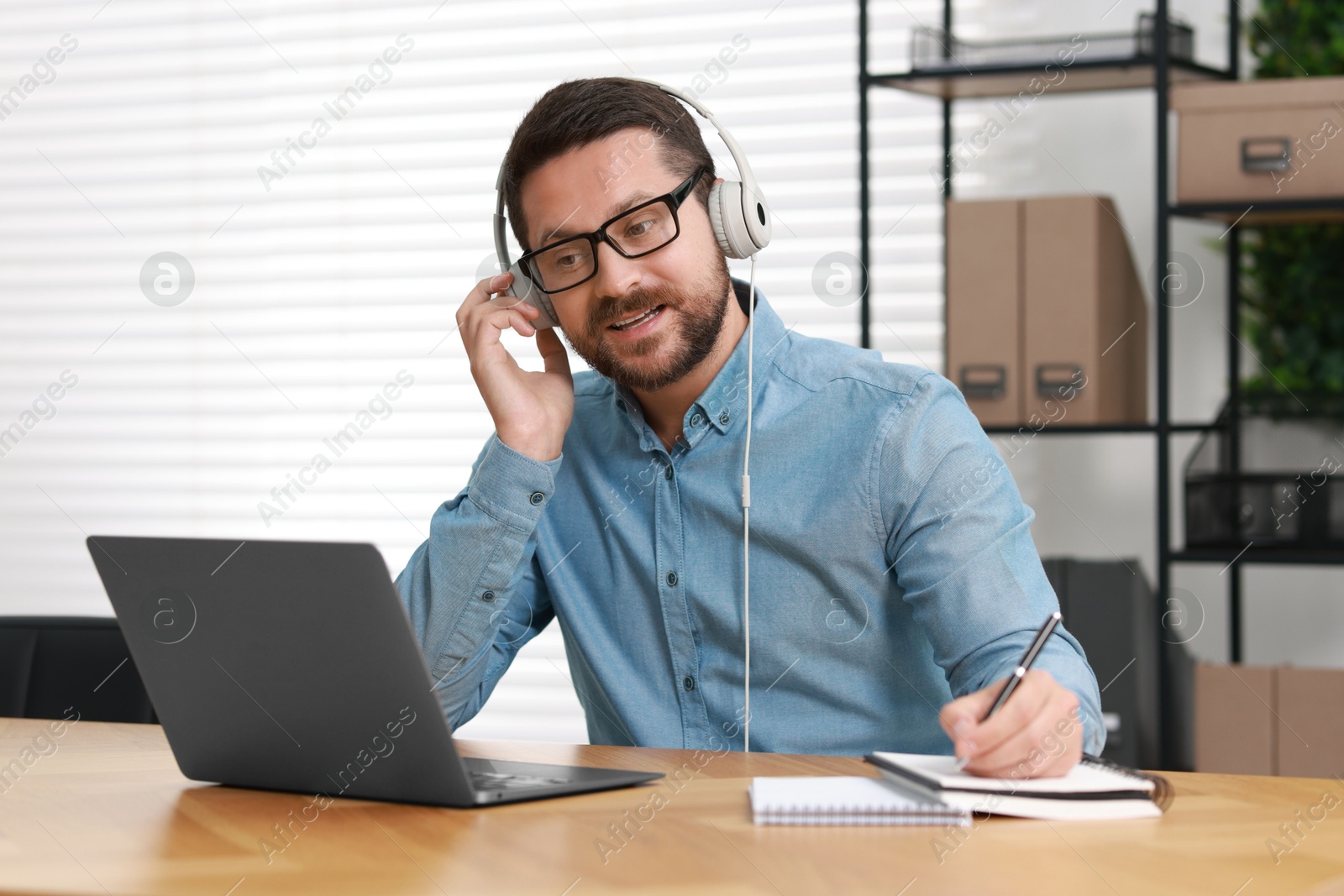 Photo of Interpreter in headphones taking notes while having video chat via laptop at wooden table indoors