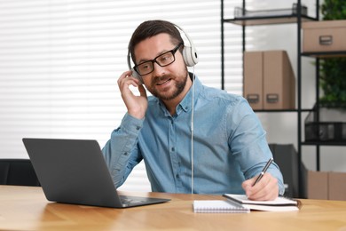 Photo of Interpreter in headphones taking notes while having video chat via laptop at wooden table indoors