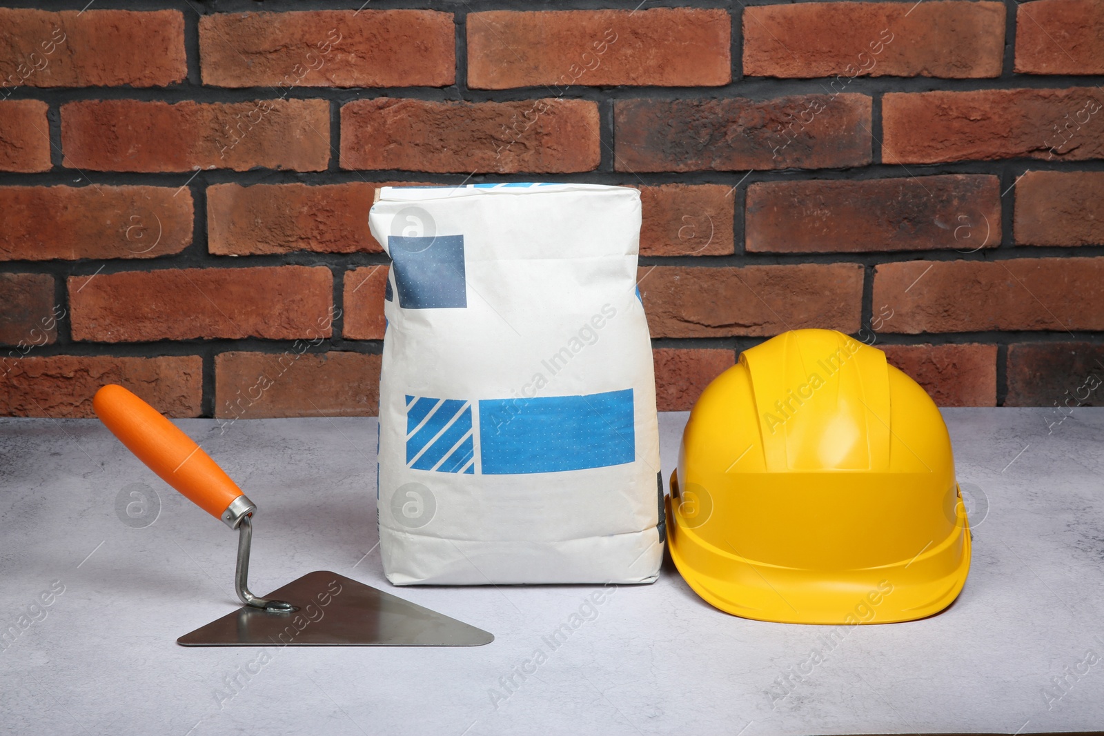 Photo of Bag of cement, hard hat and trowel on light textured table against red brick wall