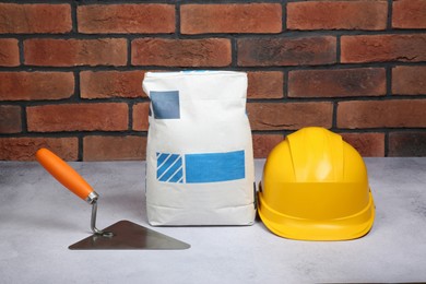 Photo of Bag of cement, hard hat and trowel on light textured table against red brick wall