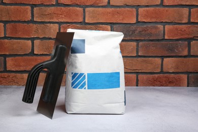 Bag of cement and plastering trowel on light textured table against red brick wall