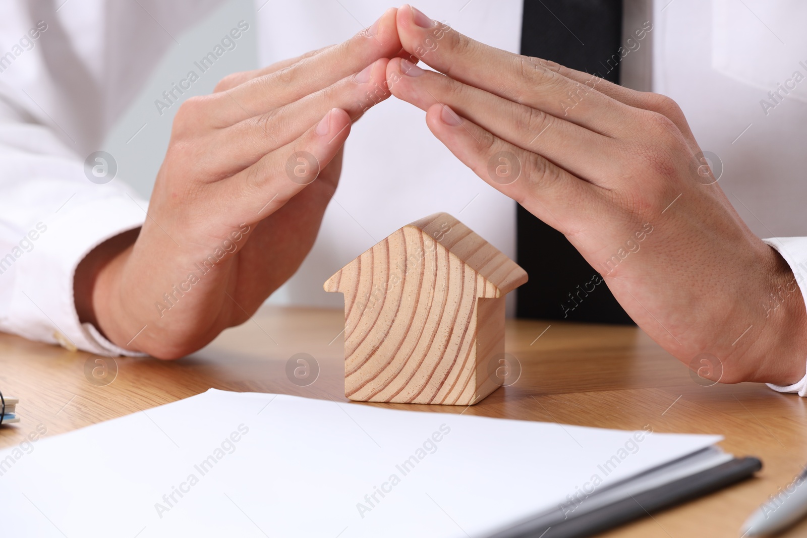 Photo of Property insurance. Real estate agent protecting house figure at wooden table, closeup