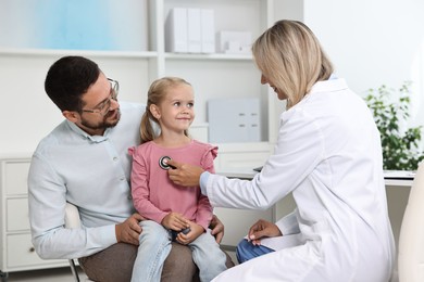 Doctor examining little girl with stethoscope and her father in hospital